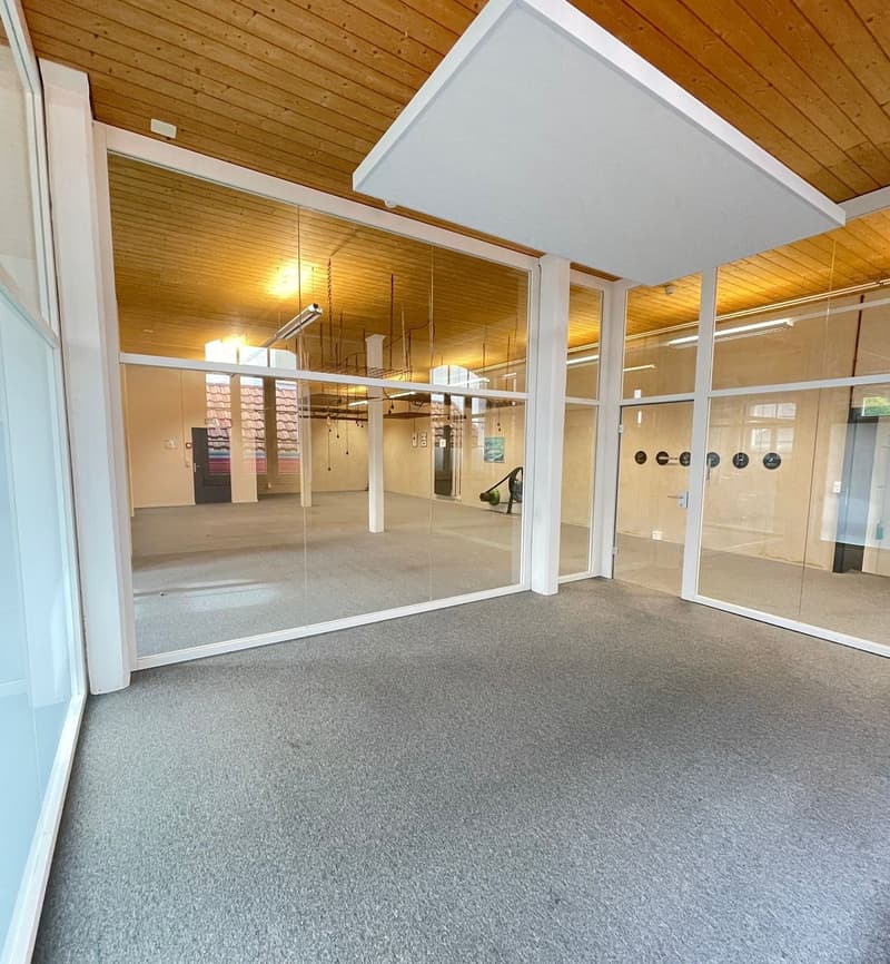 The image shows a large, open commercial space with a wooden ceiling and walls. The space has a grey carpeted floor and large glass windows that allow natural light to enter. There are various lighting fixtures and equipment visible, suggesting this could