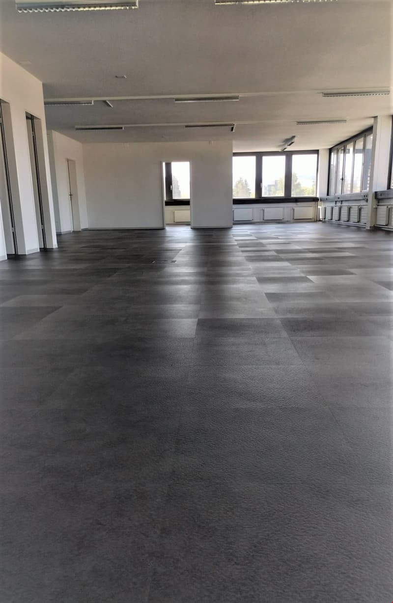 The image shows a large, open office space with a gray tiled floor and white walls. The room has several windows providing natural light, and there are various lighting fixtures installed in the ceiling. The space appears to be empty and ready for use.