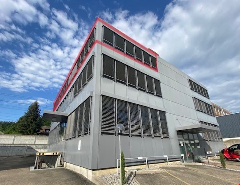 A multi-story commercial building with a gray exterior and red accents. The building has large windows and a parking area in front.