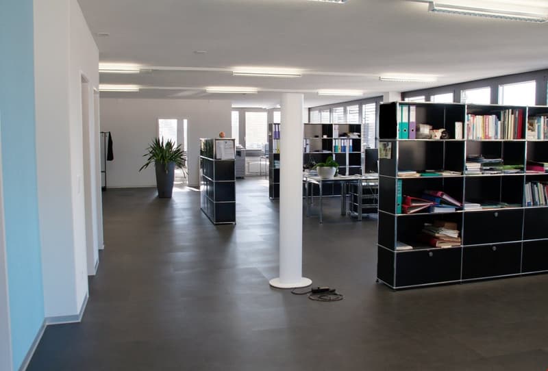 Open office layout with gray walls, hardwood floors, black shelving units, desks, and office equipment