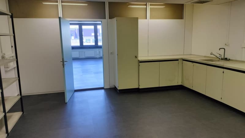 The image shows a spacious office space with a kitchenette area. The room has white walls, a large window, and a concrete floor. The kitchenette area features cabinets and a sink. The space appears to be a clean and modern office environment.