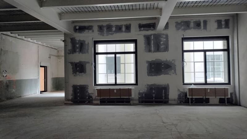 Empty room, concrete floor, unfinished walls, two large windows, door at the end of the room, ceiling with exposed beams.