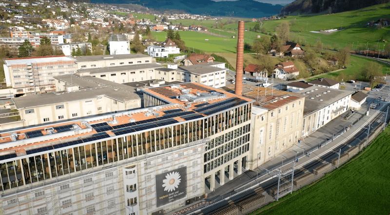 A large multi-story building complex with solar panels on the roof, surrounded by greenery and a backdrop of hills. The building features glass windows, a tall chimney, and various other architectural details. Several smaller houses and structures are vis