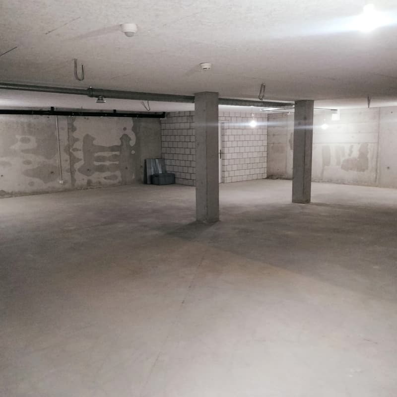 Empty parking space, concrete floor, white walls, white ceiling, white pipe on ceiling, metal pipe on wall, two lamps on ceiling
