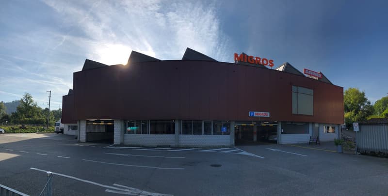 A Migros supermarket with a sloping roof, brick walls, large windows, a parking lot, and several cars parked