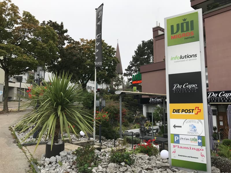 The image shows a commercial area with various businesses and shops, including a restaurant called 'Da Capo', a post office, and a Flamingo store. There are trees, plants, and a parking area visible in the scene.