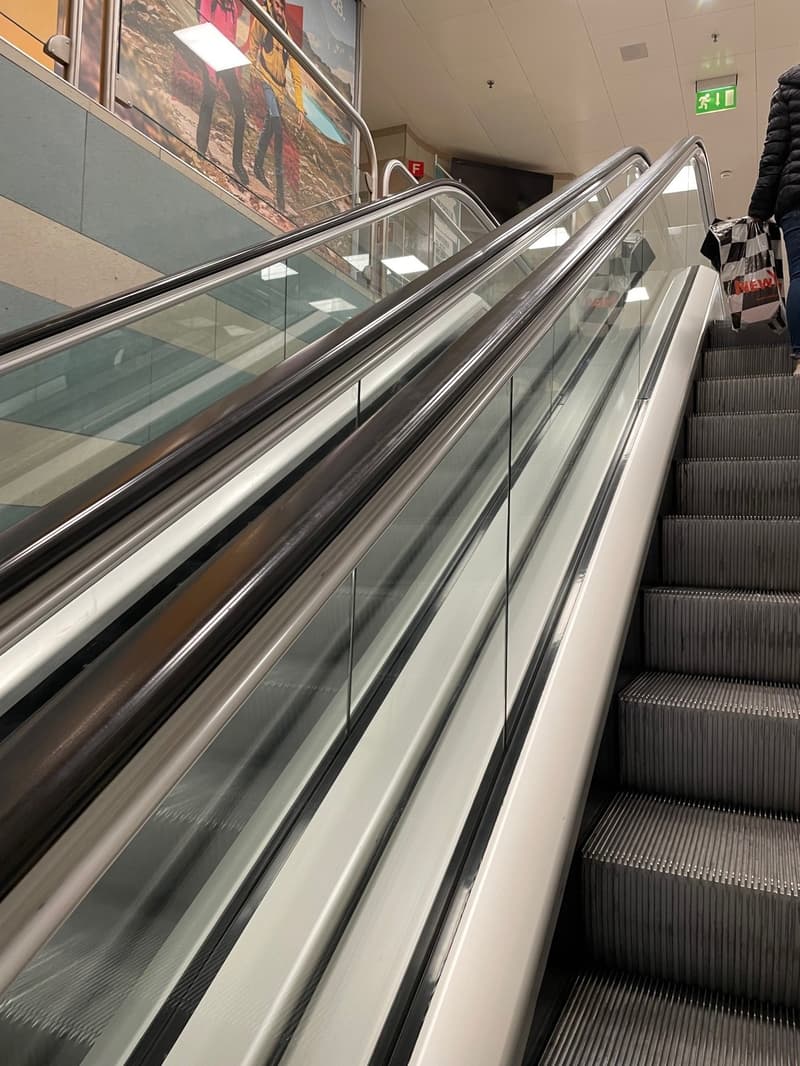 Escalator, person carrying a bag with the word NEW on it, walls with pictures, ceiling with lights