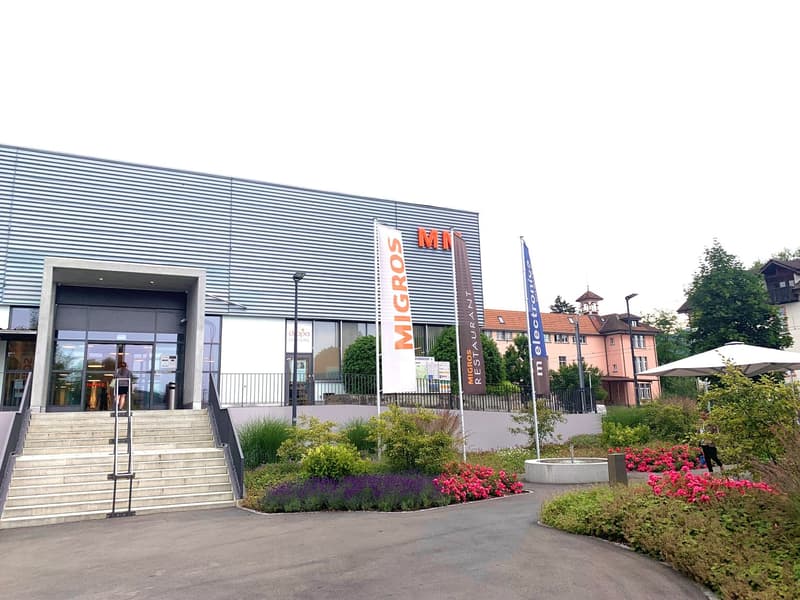 Concrete building with a large entrance and stairs leading to the entrance. There is a garden in front of the building with different plants and flowers.