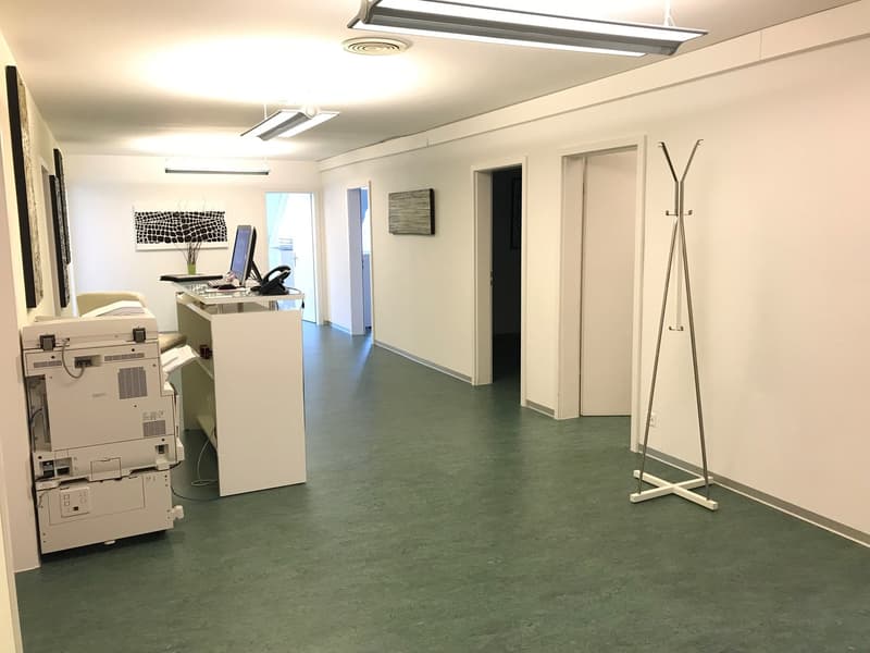 This image shows an office space with a desk, computer, and printer. The room has white walls and a gray concrete floor. There is a coat rack and a piece of artwork on the wall. The space appears to be well-lit with overhead lighting fixtures.