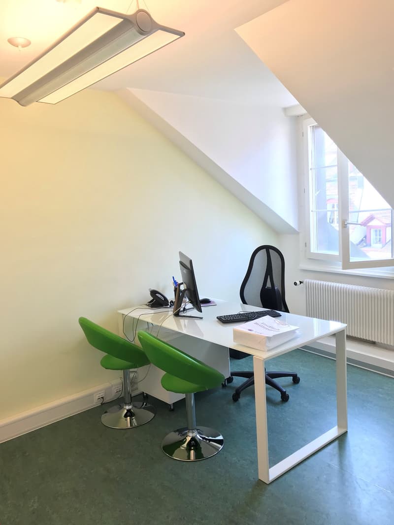 The image shows a small office space with a desk, computer, and two green swivel chairs. The room has white walls, a large window, and a ceiling-mounted light fixture. The floor appears to be a light-colored tile or laminate material.