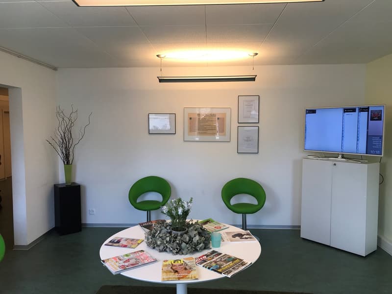 The image shows an office space with white walls, a large TV mounted on the wall, and two green chairs around a circular table with magazines and a vase of flowers. There is also a black cabinet and a tall plant in the corner of the room.