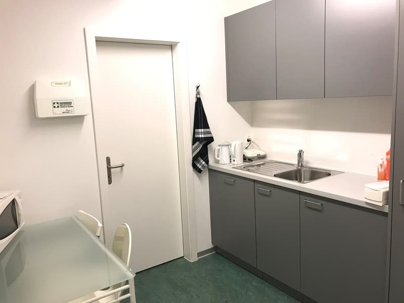 Small kitchenette with gray cabinets, sink, and appliances. There is a white door, a towel hanging on the cabinet, and a microwave on the counter.