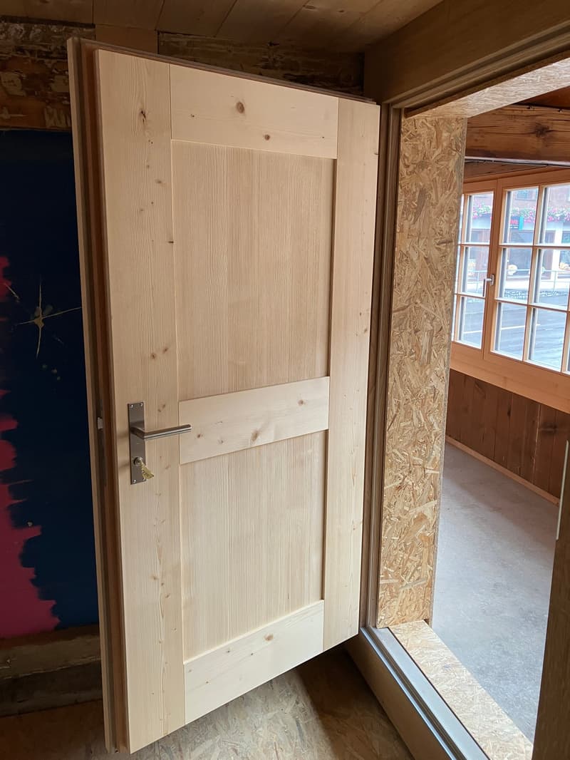 The image shows a wooden door frame and door in a room with wooden walls and floors. The door appears to be made of natural wood panels and has a simple, minimalist design. The room has a rustic, cabin-like feel with the exposed wooden construction materi