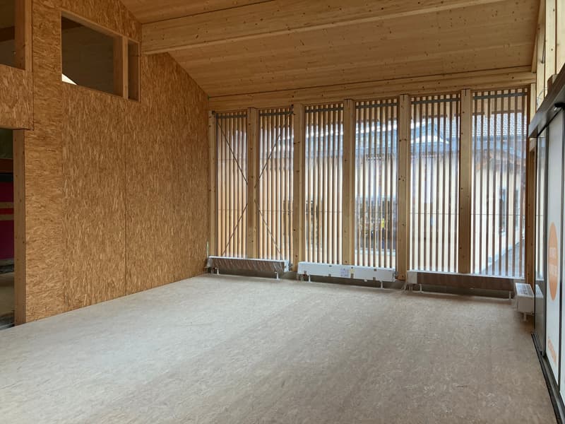 Large open space with wooden walls and ceiling, concrete floor, and large windows providing natural light. The room appears to be a living or multipurpose area.