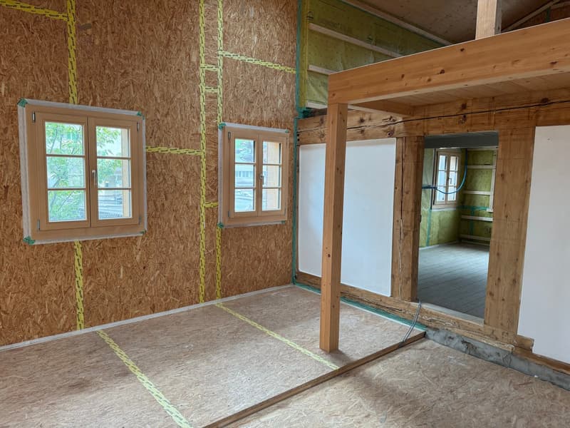 The image shows an unfinished interior space with exposed wooden framing and OSB (oriented strand board) walls. There are two windows with wooden frames, and the floor is covered with plywood or OSB panels. The room appears to be in the process of constru