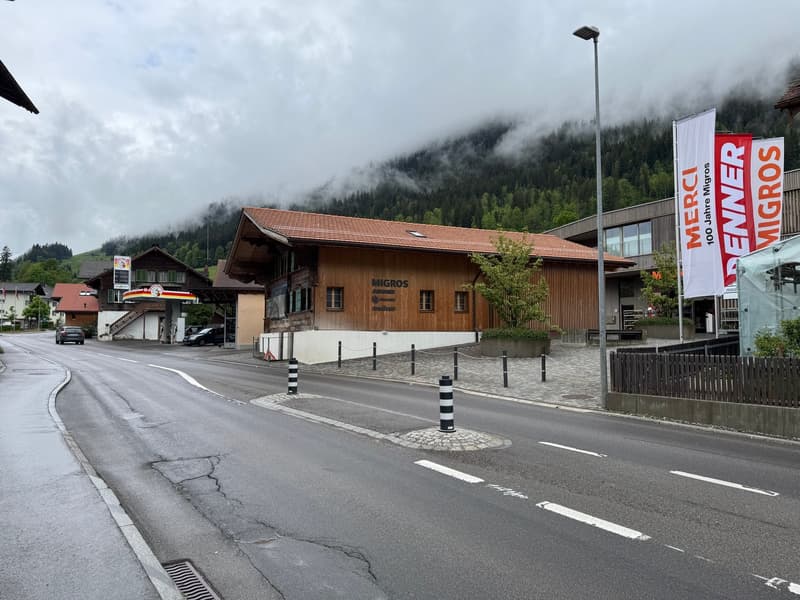 A two-story wooden building with the Migros logo, located in a mountainous area with fog in the background. There is a parking area in front of the building and a road with road markings.