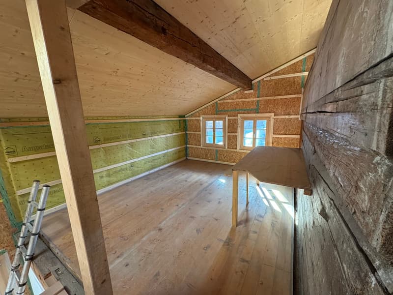 The image shows an unfinished attic or loft space with exposed wooden beams and walls. The floor is made of wooden planks, and there are two small windows that let in natural light. The space appears to be in the process of renovation or construction, wit
