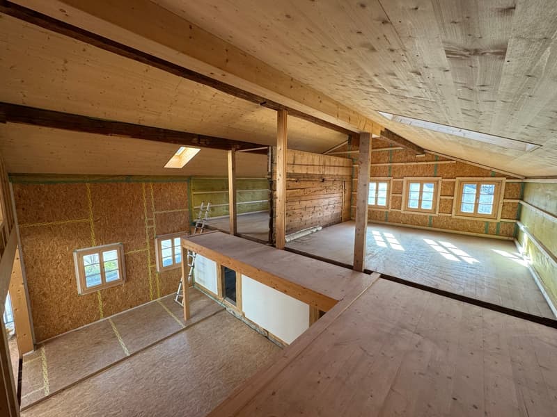 The image shows an unfinished attic space with exposed wooden beams and trusses, plywood flooring, and several windows providing natural light. The space appears to be in the process of renovation or construction, with ladders and other construction mater