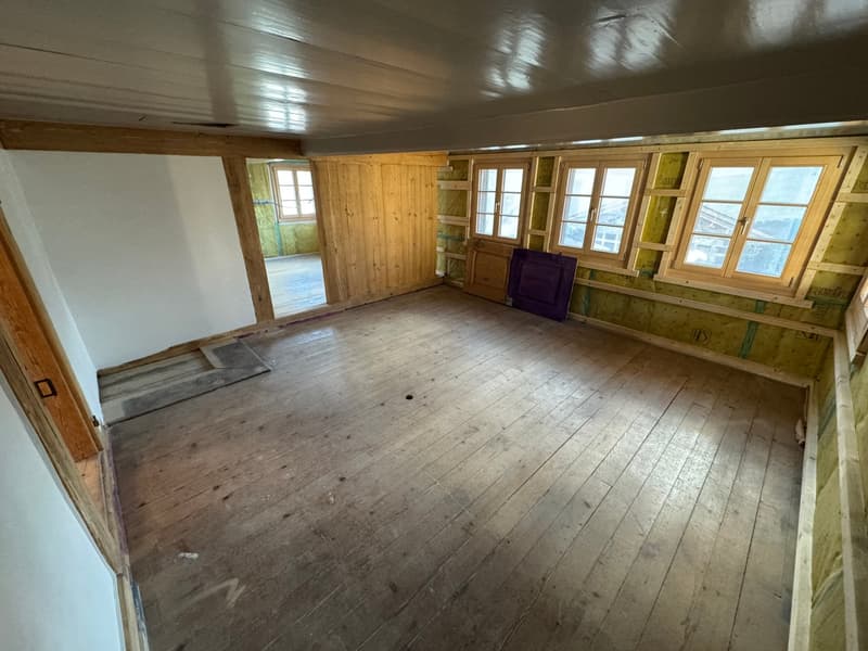 This image shows a large, open room with wooden walls and floors. The room appears to be in a state of renovation or construction, with some unfinished areas and a lack of furniture or decor. The room has several windows that provide natural light, and th