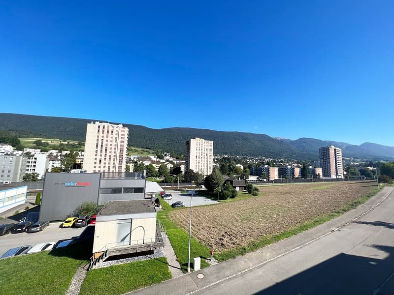 The image shows a cityscape with several high-rise apartment buildings and a large open field in the foreground. The buildings appear to be located in a mountainous area, with a clear blue sky overhead. There is a parking lot and a small building with the