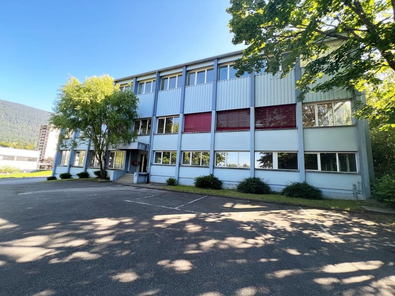 A multi-story commercial or institutional building with a modern, modular design. The building has large windows, a gray exterior, and is surrounded by trees and a paved parking lot.