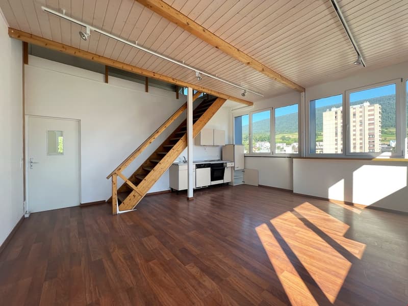 Open-concept studio apartment with wooden ceiling beams, hardwood flooring, large windows providing natural light, and a staircase leading to a loft or upper level. The kitchen area is visible with appliances such as a stove and refrigerator.