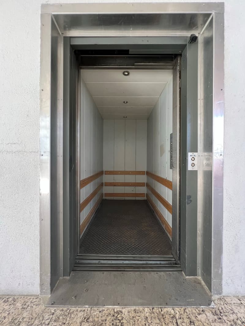 The image shows the interior of an elevator. The elevator has a metal frame and a diamond-patterned metal floor. There are wooden steps leading up to the elevator platform. The walls of the elevator are white and there is a control panel visible on the si