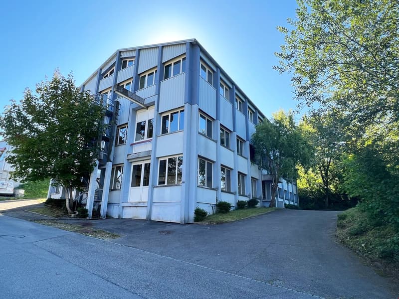 A multi-story commercial or office building with a modern, industrial-style design. The building has a gray exterior with large windows and is surrounded by trees and greenery.
