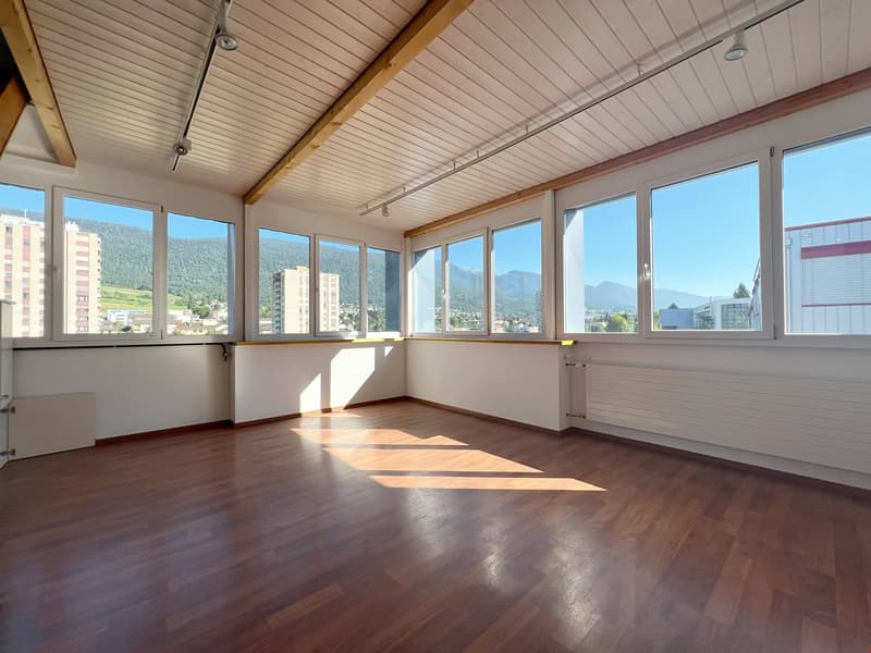 Spacious room with large windows offering a scenic view of the surrounding mountains and cityscape. The room has a wooden floor and a paneled ceiling with exposed wooden beams. The room appears to be a living or office space with ample natural light.