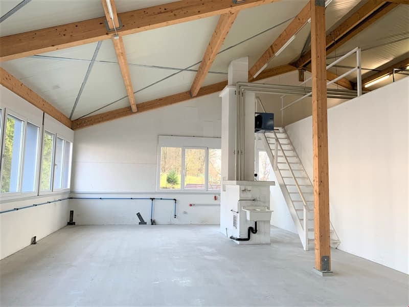 This image shows the interior of a building under construction or renovation. The space has a high ceiling with exposed wooden beams and trusses, and the floor appears to be concrete. There is a small sink or utility fixture installed, and a ladder leadin