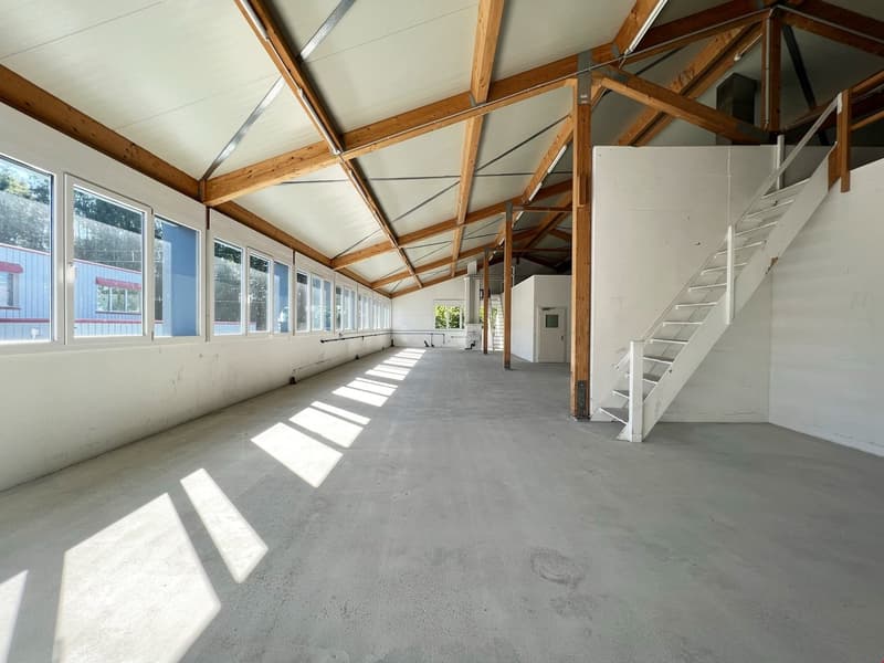 The image shows a large open commercial space with a high ceiling and exposed wooden beams. The space has large windows along the walls, allowing natural light to flood in. The floor is a concrete slab, and there is a staircase leading to a second level.