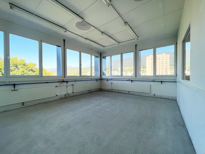 Large open room with concrete floor, white walls, and large windows providing natural light. The room has a suspended ceiling with air vents and lighting fixtures.