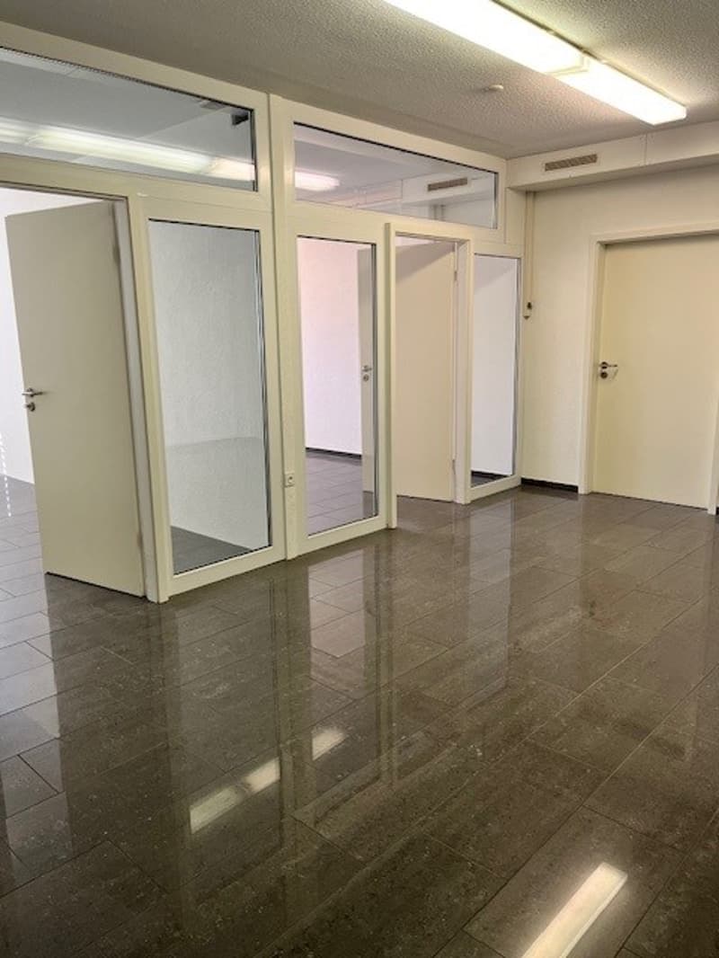 The image shows an empty office space with a tiled floor and glass-paneled doors. The room has a clean, modern appearance with neutral-colored walls and overhead lighting.