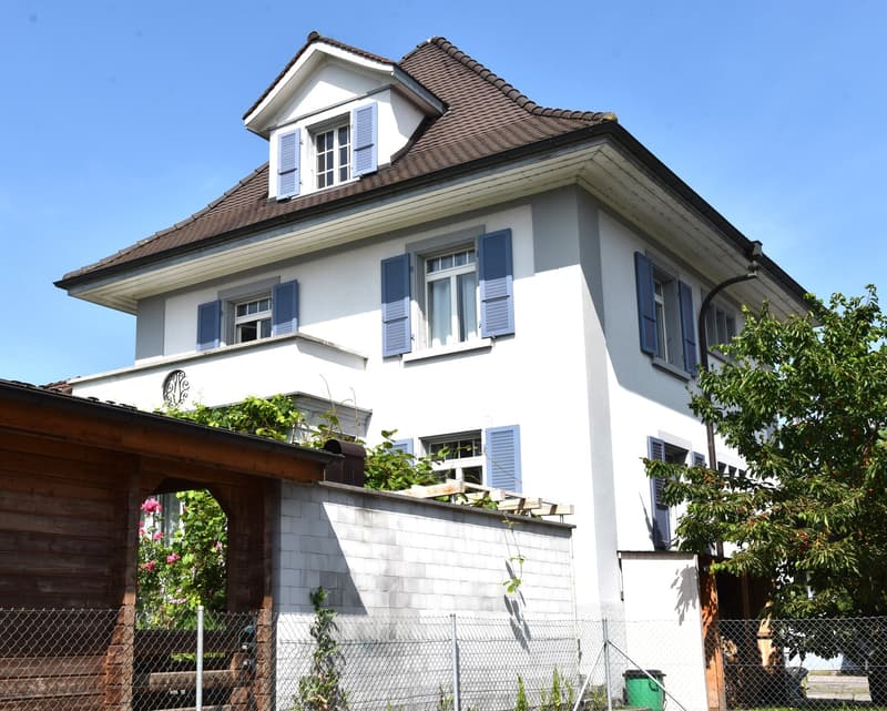 2-story house, white exterior, brown roof, multiple windows with blue shutters, front balcony, side wall covered with vines, fenced garden