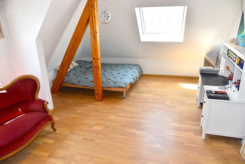 A small bedroom in the attic, equipped with a single bed, a red armchair, and a desk with shelves.