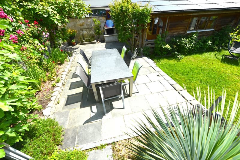 Dining area with table and chairs, green plants, small garden with brick and stone elements, exterior fireplace, and wooden house with a solar panel on the roof.