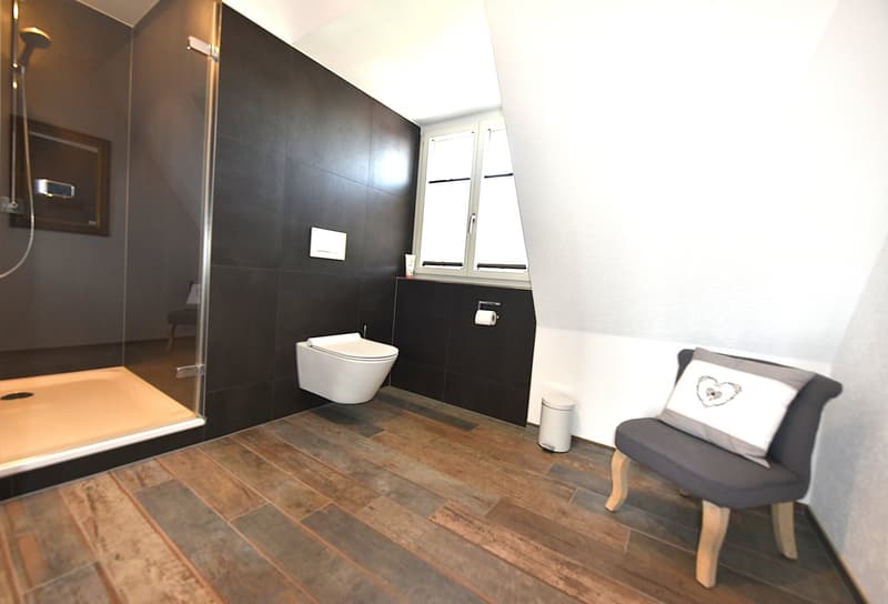 Bathroom: glass shower, toilet, tiled floor, window, chair, trash bin, toilet paper roll