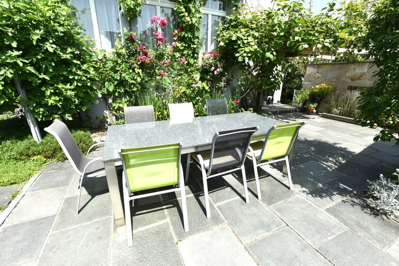 Table and chairs in the garden area, covered with climbing plants, wall, trees, and grass.