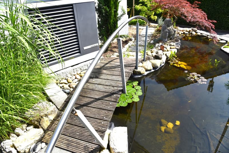Wooden walkway leading over a pond, surrounded by rocks and plants, a small statue, and a curved metal railing