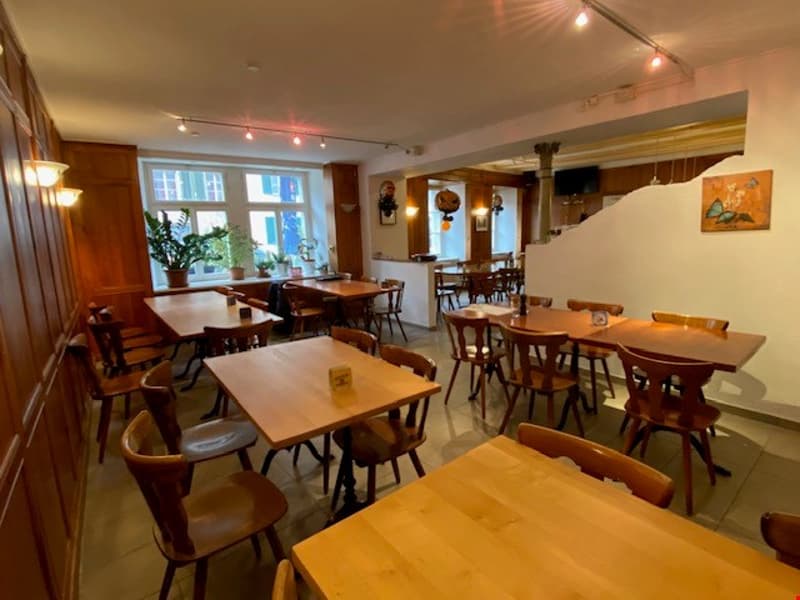 Modern restaurant interior with wooden chairs and tables, warm lighting, potted plants by the windows, decorative artwork on the wall, a white ceiling with integrated lighting
