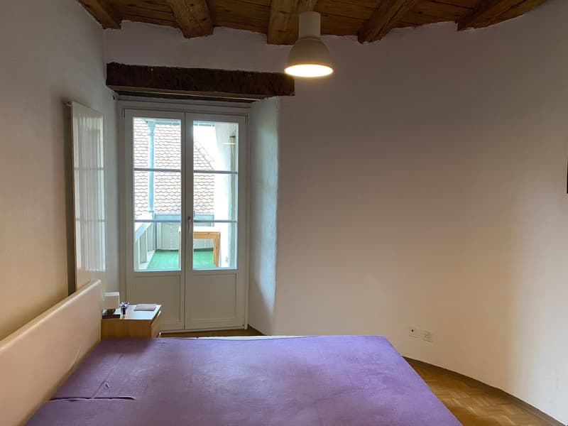 Bedroom with wooden ceiling beams, white walls, large window with balcony access, and a purple bedspread on the bed.