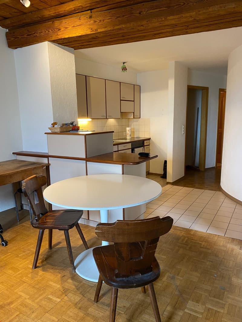 The image shows a kitchen area with wooden ceiling beams, white cabinets, a round dining table, and wooden chairs. There is a kitchen counter with a built-in dishwasher visible.