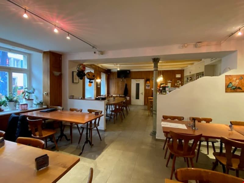 The image shows a cozy and rustic-style restaurant or cafe interior. The space features wooden tables and chairs, exposed brick walls, and track lighting. There appears to be a kitchen or bar area visible in the background. The visible features include a