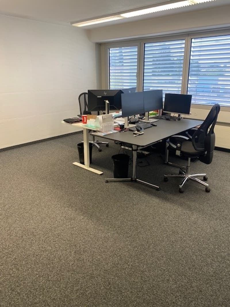 The image shows an office space with multiple desks, computers, and office chairs. The room has large windows and appears to be on a raised ground floor.