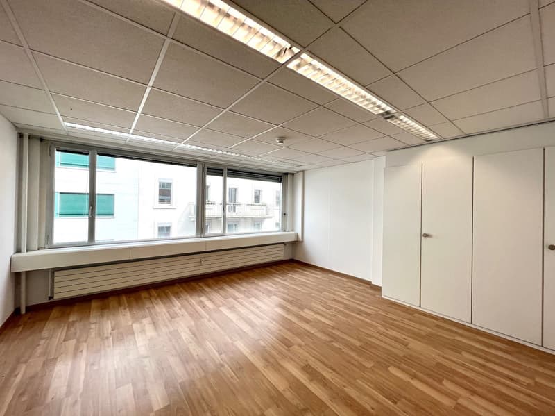 An empty room with wooden floors and white walls. Large windows with white window sill and green shutters. White built-in cabinets and a white ceiling with integrated fluorescent lighting.