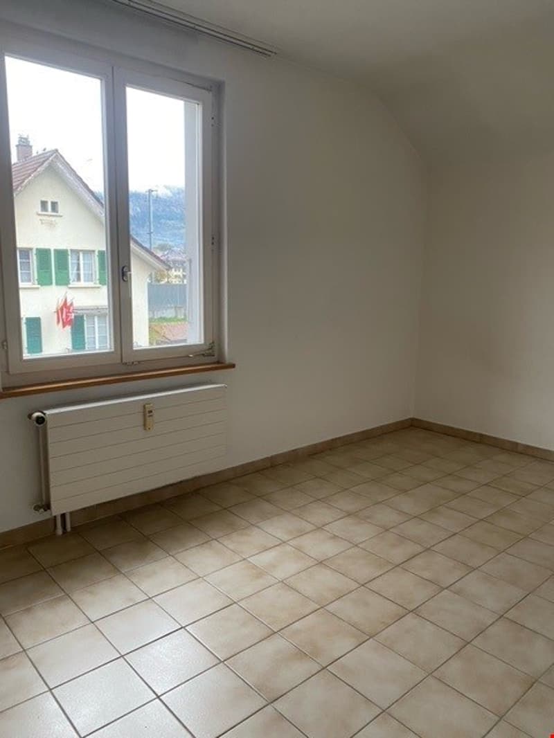 Empty room with white walls, beige tiles, large window, and a white radiator