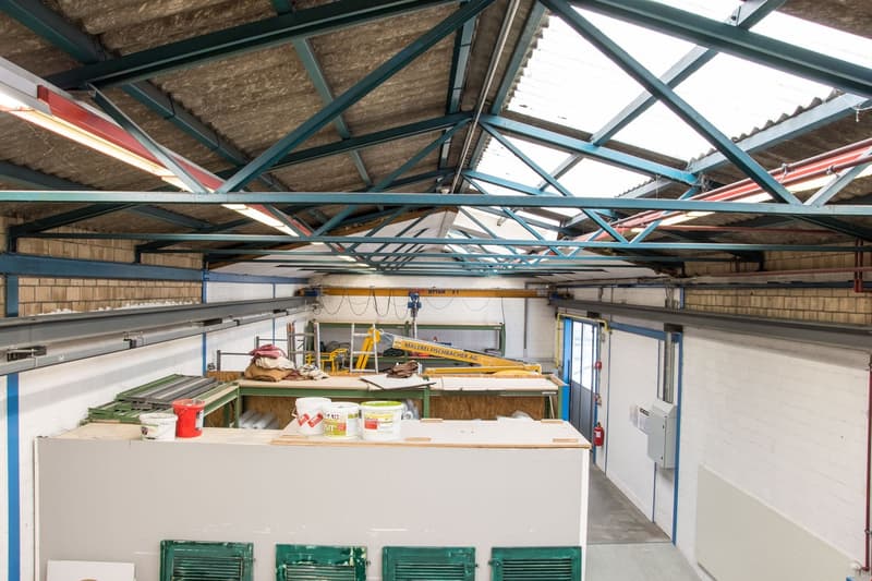 Industrial workshop with metal roofing, large windows, white walls, several workbenches, metal storage units, tools, buckets, ladder, metal cranes, extinguisher, open door
