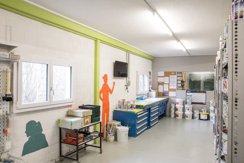 Large room with white walls, green trim, large windows, industrial shelves, desk, shelves filled with paint cans, monitor, tools.
