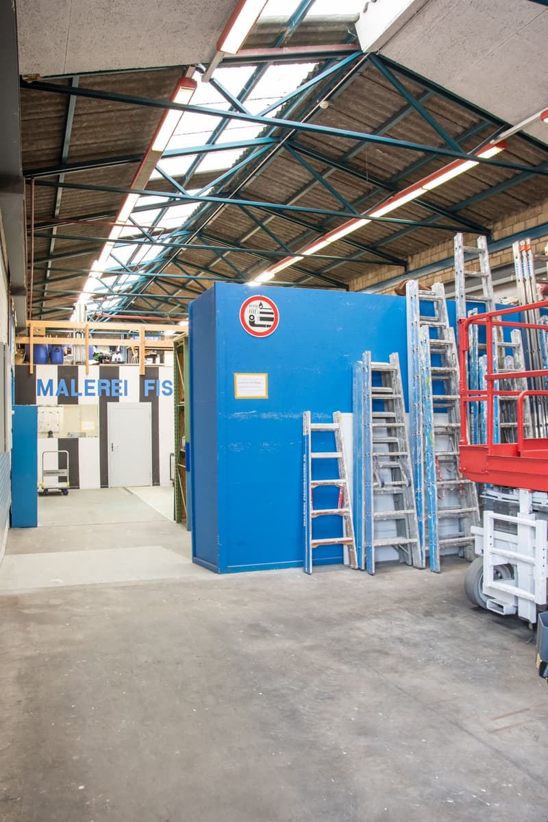 Warehouse with an open interior space, featuring a high ceiling with exposed metal beams and industrial lighting. A large blue storage unit is against one wall, with shelves and storage containers on top. Several ladders and a red forklift are also presen