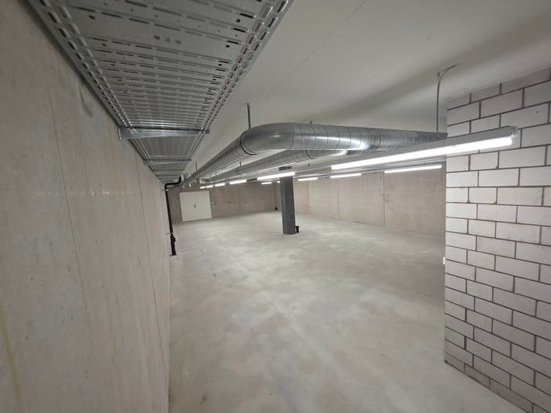 Empty parking space, gray floor, gray walls, white ceiling, metal ducts, metal pipes, white door, lighting